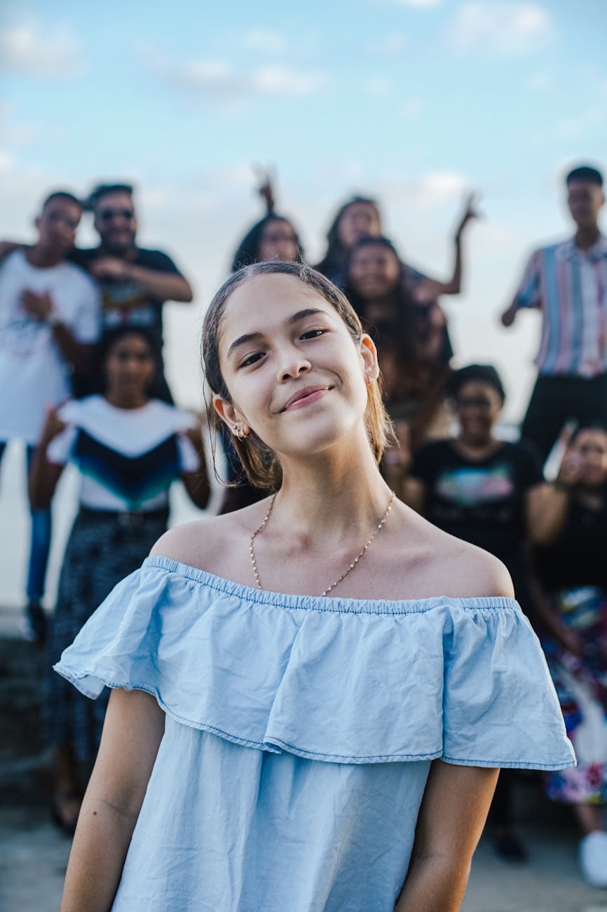 Alt text: "A young woman in a blue off-the-shoulder top smiles gently at the camera, with a diverse group of cheerful people in casual attire blurred in the background, representing the vibrant community of the SEO Surfers group on Facebook.
